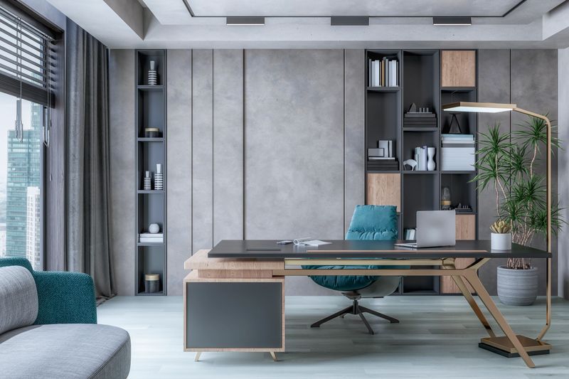 A director's office interior with a work desk, a chair and decoration (a laptop, phone, pens, a workbook and a succulent plant) on gray hardwood floor in front of a concrete gray wall, a library (shelves with books and sculptures) and copy space. A large potted plant dracaena marginata, a high golden electric lamp, a sofa and large windows with cityscape view (skyscrapers), blinds and gray curtains aside . Built-in recessed ceiling gray lights (reflectors) with grooves and channels. A 3D rendered image.