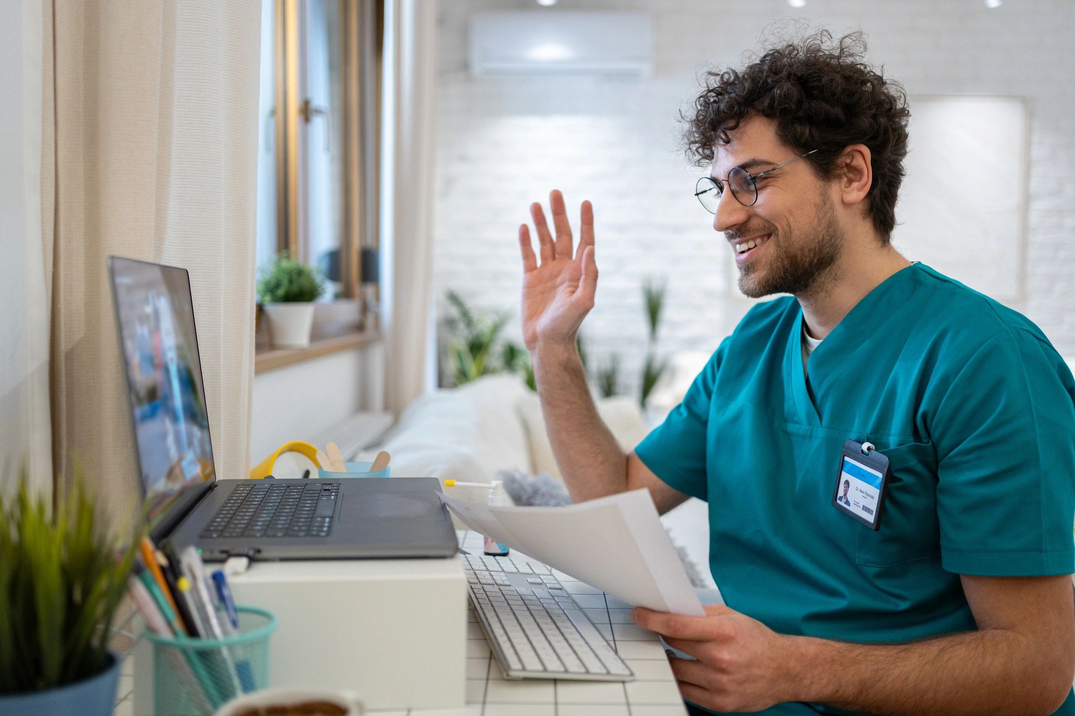 Doctor in scrubs smiling and waving during a video call.
