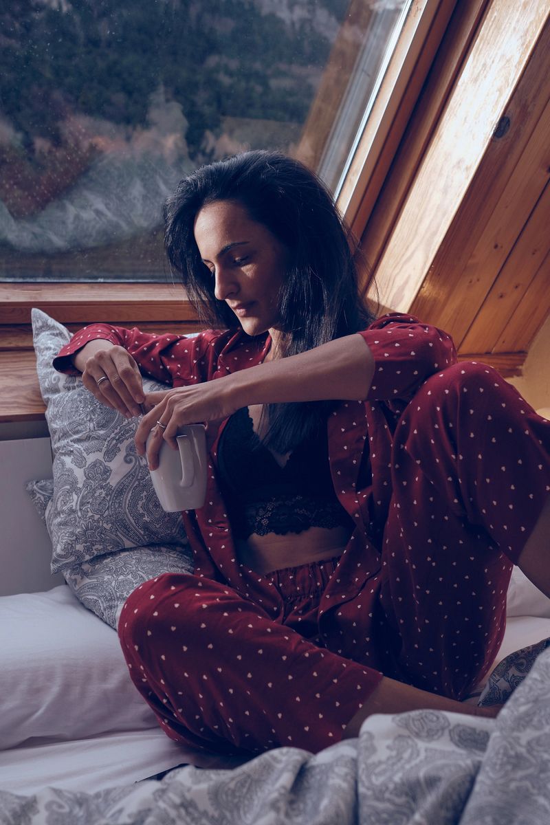 Tranquil female in nightwear sitting with cup of drink on bed in house in mountains and relaxing