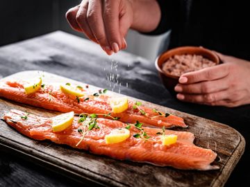 Hand sprinkling salt on lemon-garnished salmon fillets on a wooden board.