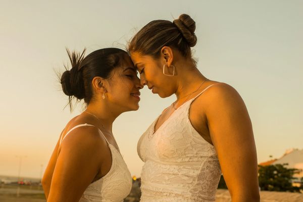 Two woman in their white bride dresses standing face to face in front of a sunset.