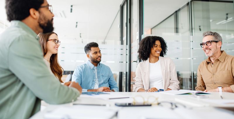 A diverse group of professionals is engaged in a lively discussion around a sleek, modern conference table in a well-lit office, reflecting a collaborative and inclusive work environment.