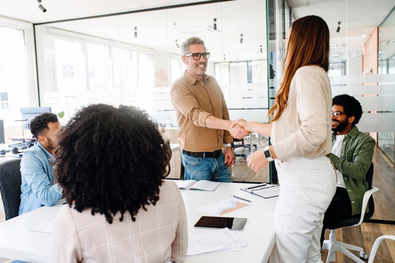 Mature grey-haired man in glasses and a joyous expression shakes hands with a young woman, creating an atmosphere of warm collaboration and team success. Diverse team involved meeting