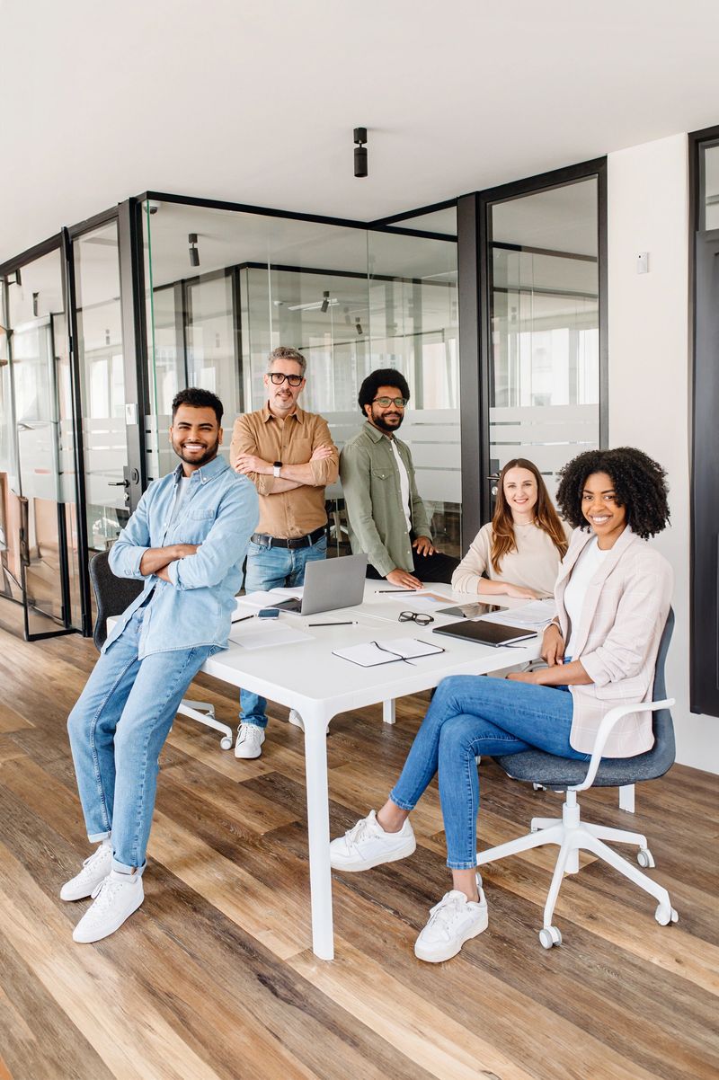 A vibrant team gathers around a minimalist white table in their bright office, embodying the energy and collaboration of a modern, dynamic workplace, diverse ages and ethnic collaborate together