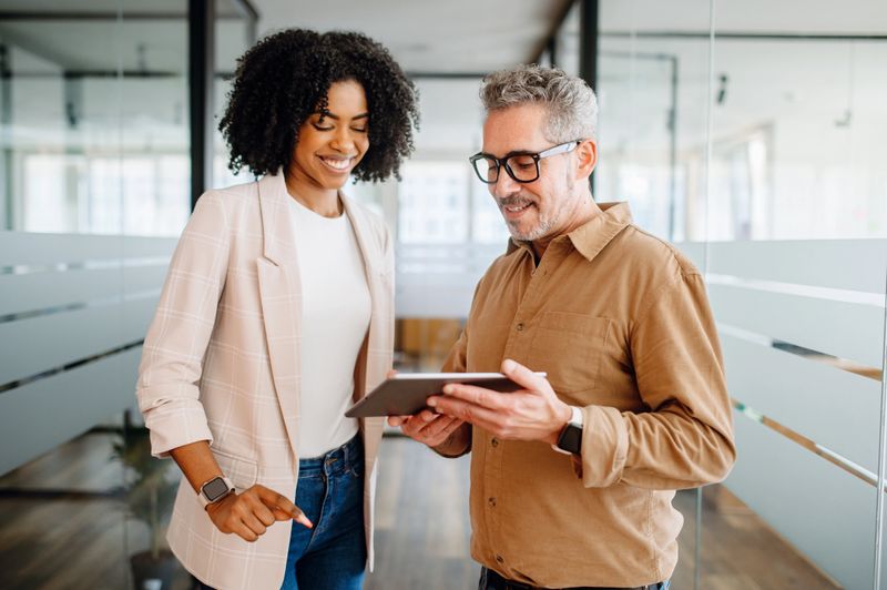 A businesswoman and a senior male colleague collaboratively review content on a tablet in a bright office space, reflecting a multigenerational workplace where experience meets new technology