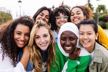 A diverse group of seven young women happily posing together outdoors.