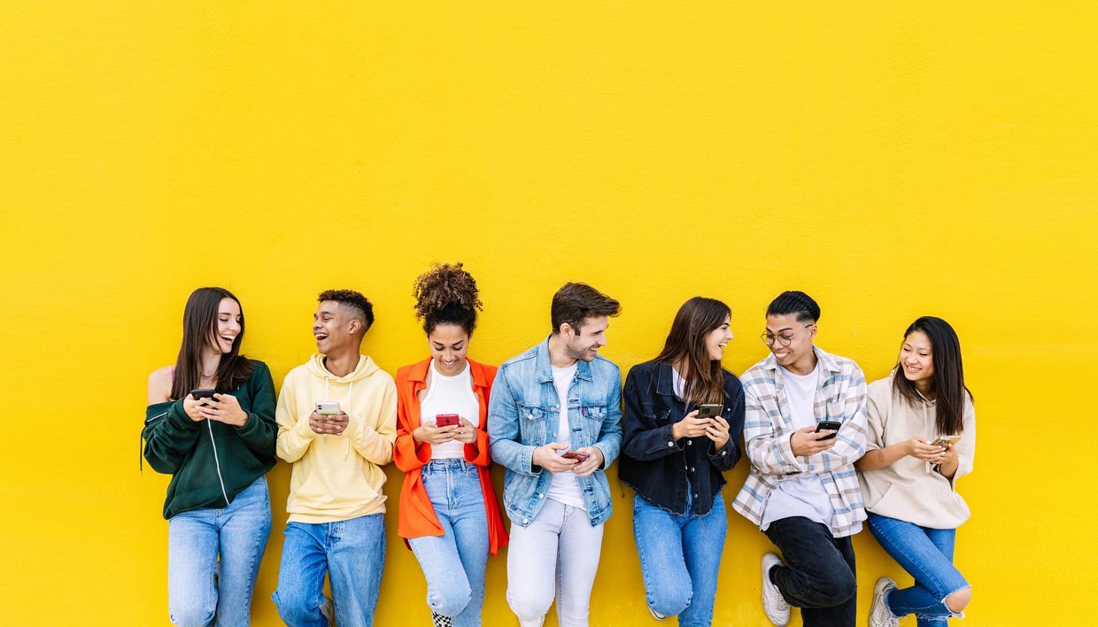 A group of diverse friends smiling and using smartphones against a yellow wall.