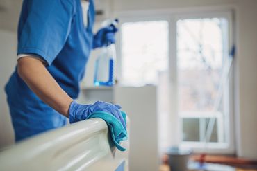 Person in blue scrubs cleaning a hospital bed rail with a cloth and spray bottle.