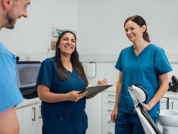 Three dental professionals in blue scrubs smiling and discussing in a clinic.