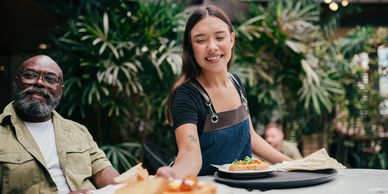 Smiling waitress serving food to a customer at a lively café with greenery.