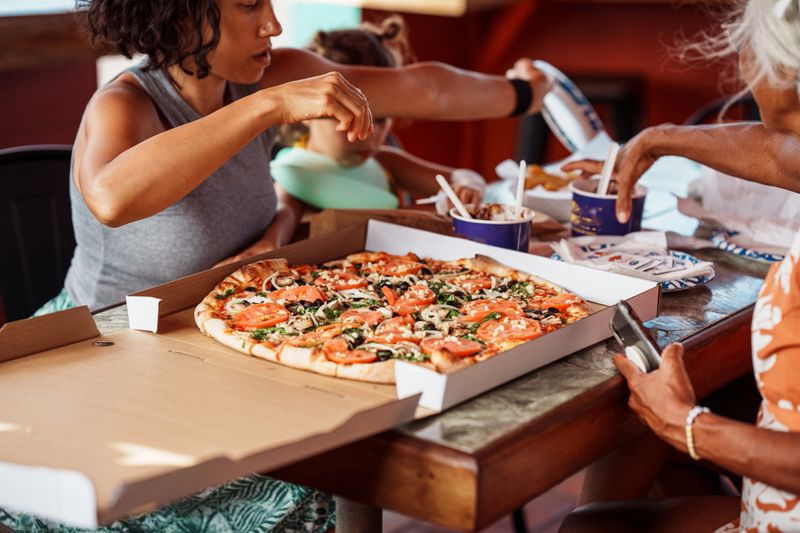 Cropped view of a multiracial, multi-generation family seated around a table and eating take-out pizza together, with selective focus on the pizza.