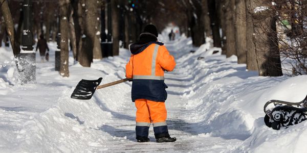 A worker in bright orange clearing snow from a path in a snowy park.