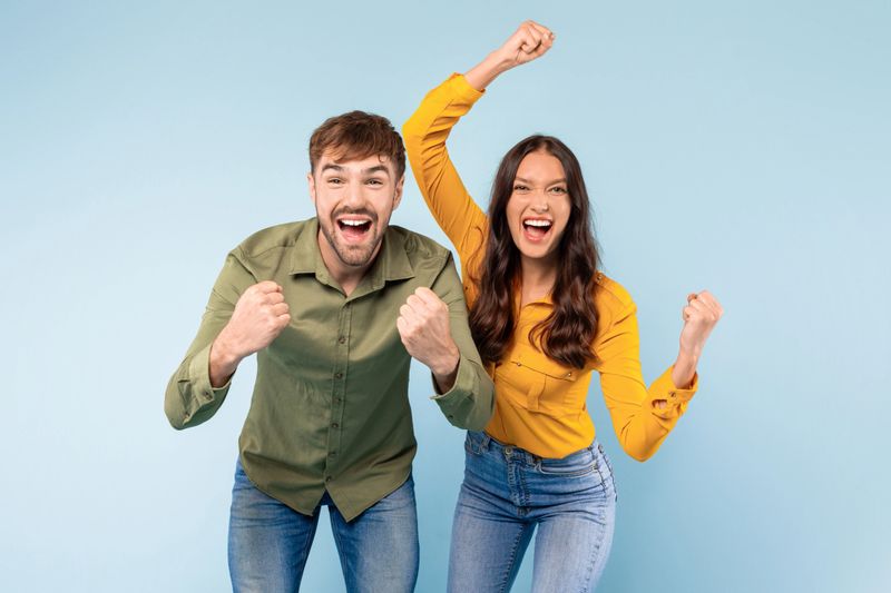 Exuberant man and woman cheerfully celebrating a victory, punching the air with raised fists, against a vibrant blue backdrop, looking at camera