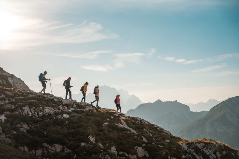 Equipped with backpacks and hiking poles, diverse friends laugh and enjoy themselves during a mountain hike, surrounded by stunning views.