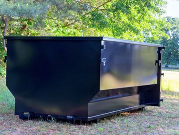Large black metal dumpster placed on grassy ground near trees.