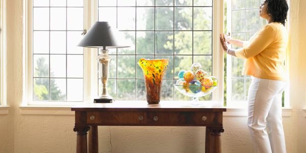 Woman opening a sunny window behind a table with decorative glass items.