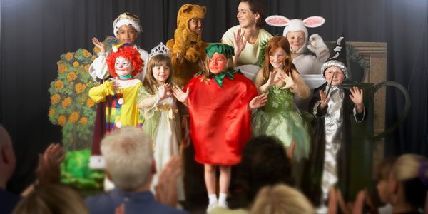 Children in colorful costumes take a bow on stage after a play, receiving applause from the audience.