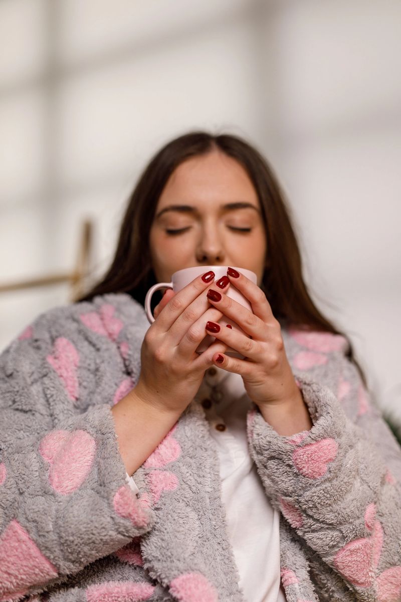 Portrait of happy young woman sitting at her home office desk, enjoying the first cup of coffee in a cozy bathrobe and pjs.