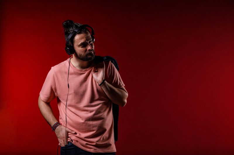 Copy space shot of cool mid adult hipster standing against bright red background, holding his jacket on the shoulder and listening to music via headphones. He is looking away, smiling and contemplating.