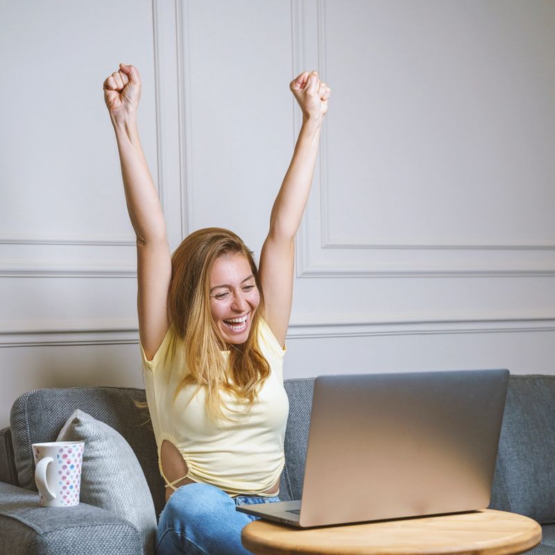 Woman working from home online while using a laptop computer and celebrating with arms up