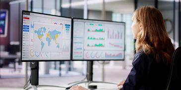 Woman analyzing global data on dual monitors in a modern office.