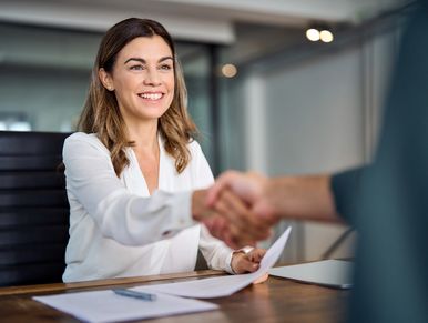 Smiling lady shaking hands at desk with papers in her other hand