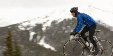 Cyclist in blue jacket riding fast on a mountain road with snowy peaks in the background.