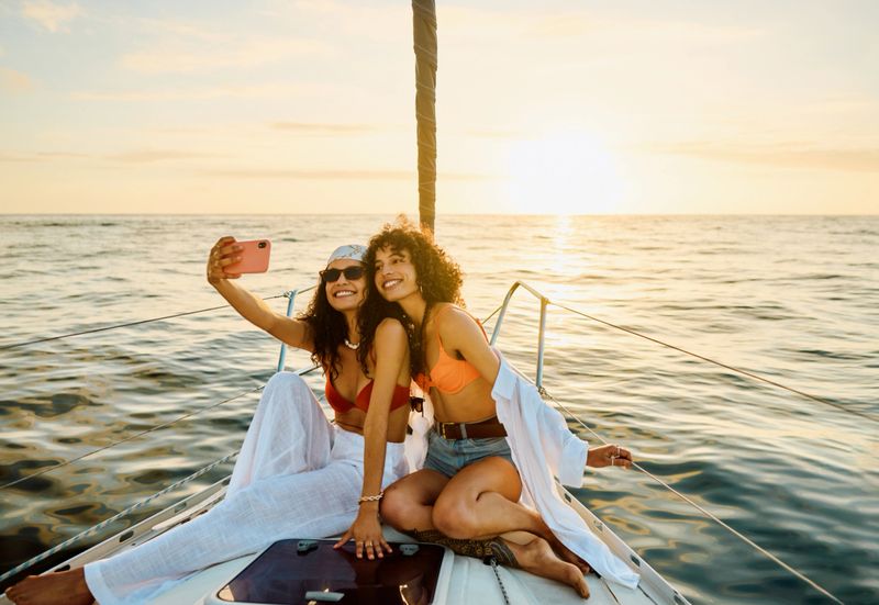 Two exuberant women in summer attire take a selfie on a yacht. With the serene ocean as their backdrop, they capture the essence of their sea adventure at sunset, complete with casual summer dresses and radiant smiles.