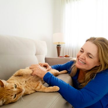 A smiling woman plays with a relaxed orange tabby cat on a beige couch.