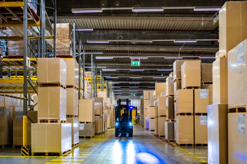 Warehouse worker driving forklift in aisle illuminated with headlights, wide shot