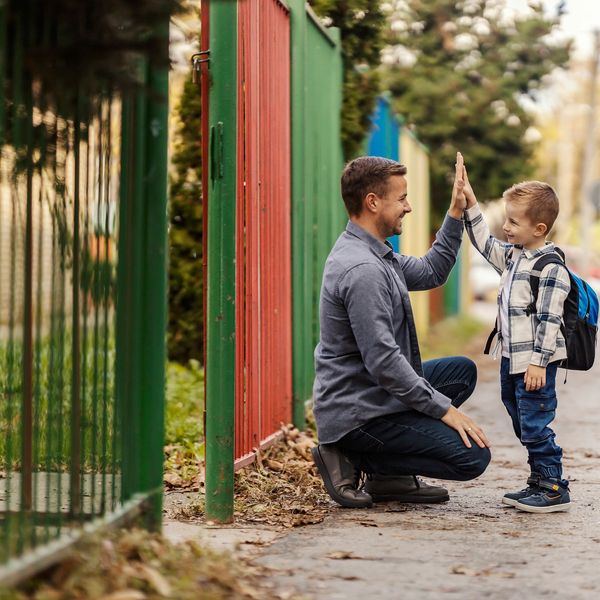 A father and son share a joyful high-five outside near a colorful fence.