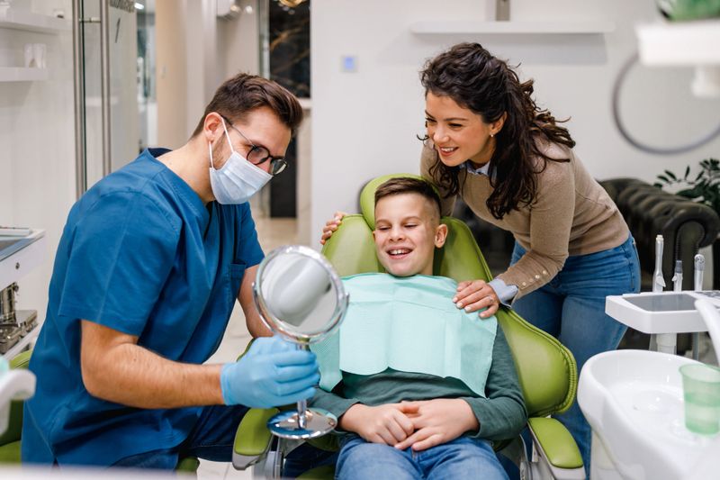 Dentist in mask and pleased mother chat while boy admires repaired teeth in mirror