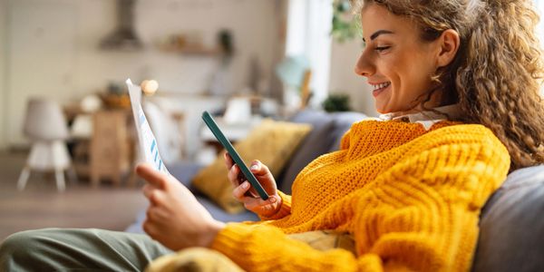 Young woman in a cozy sweater using phone and reading paper at home.