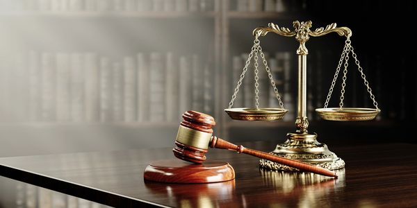 Justice balance scales and court gavel sitting on table with books on shelf in background. 