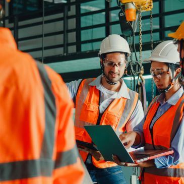 Engineers in safety gear discussing work on a laptop at an industrial site.