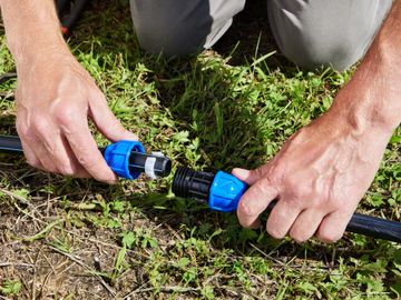 Hands connecting two black irrigation pipes with blue connectors on green grass.