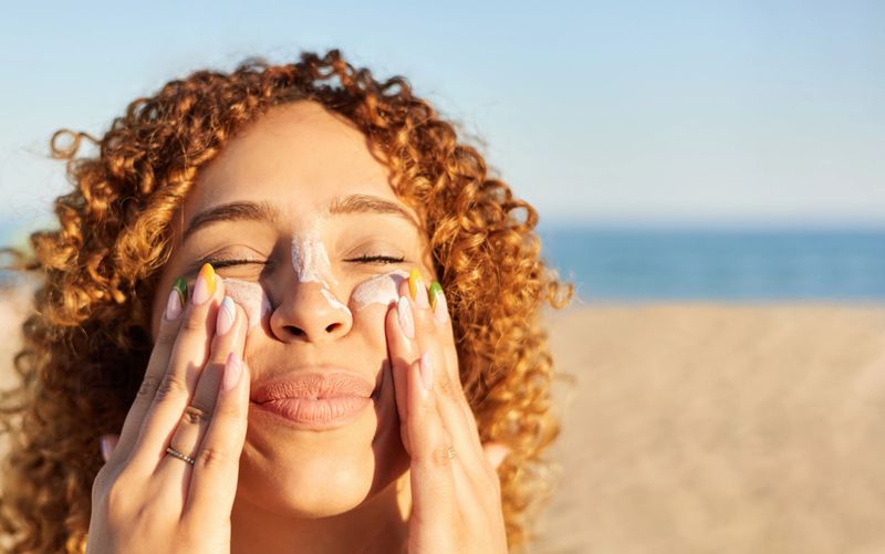 Young latin woman applying sunscreen to her face on the beach in a summer sunset. Cheerful gesture with her eyes closed
