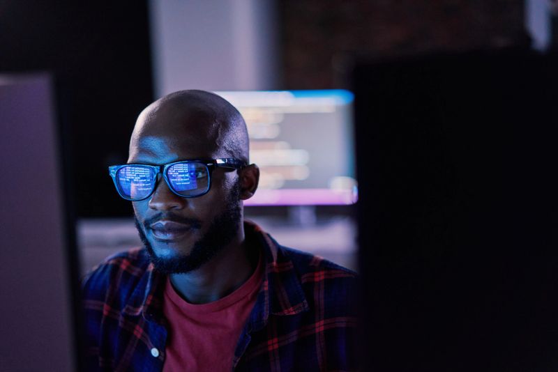 Cyber security, black man and code reflection in eyewear, hacking and software update in office. African American male employee, programmer or IT specialist with glasses, focus and cloud computing