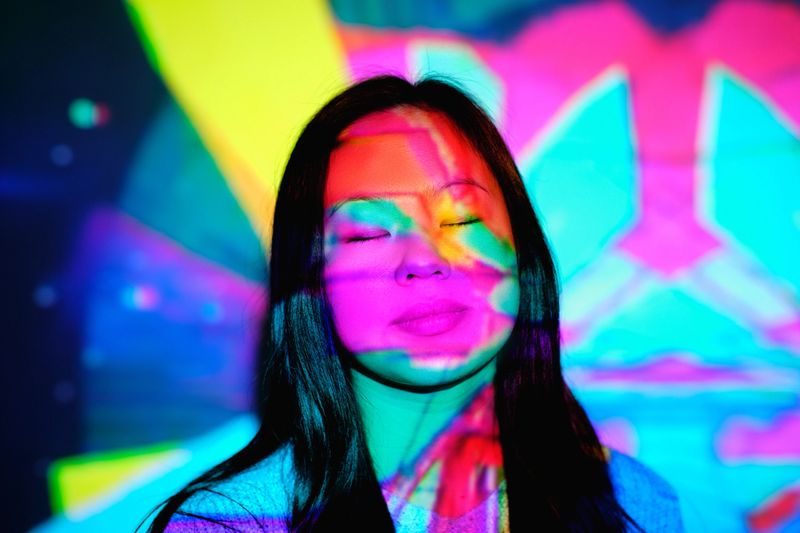 Charming Asian female with long hair standing with closed eyes under bright glowing neon lights on face in stylish studio