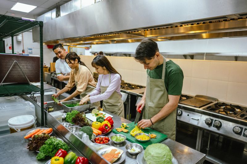 Multiracial Group of Adult Students in Aprons Slicing and Seasoning Ingredients for Dish Prepared During Team Cooking Class - Professional Cooking School