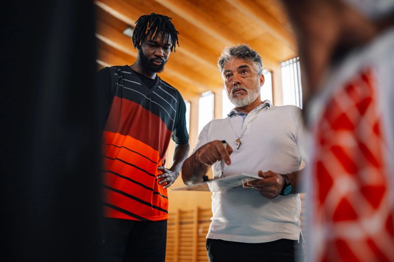 A professional senior basketball trainer explaining game plan to his multicultural team while standing on court indoors on training. A coach is coaching while interracial players are listening.