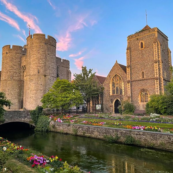 Historic stone castle and church by a flower-lined river at sunset.