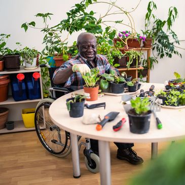 A man in a wheelchair gardening indoors with potted plants and tools.