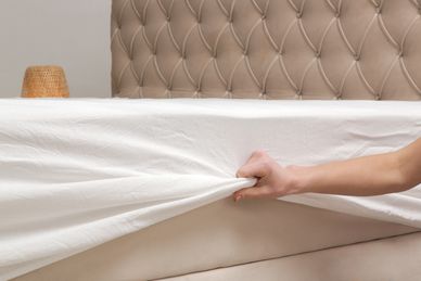 Hand pulling a white bedsheet off a beige tufted bed.