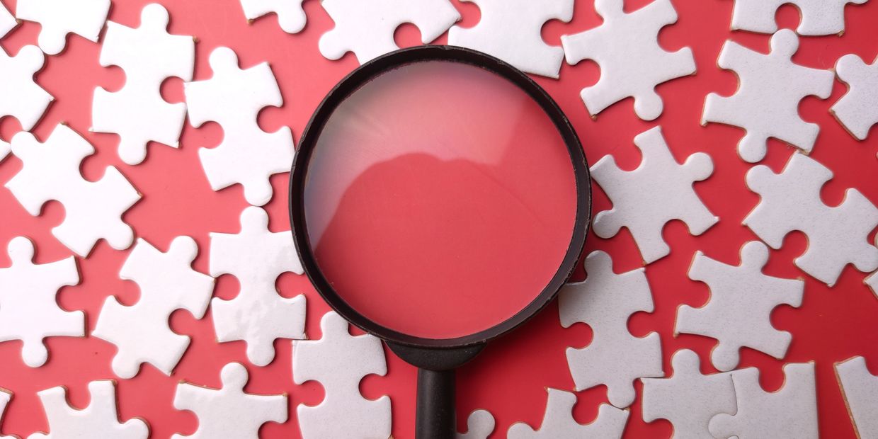 Magnifying glass focusing on white puzzle pieces scattered on a red surface.