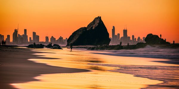 Sunset over a beach with rock formations and city skyline in the background.