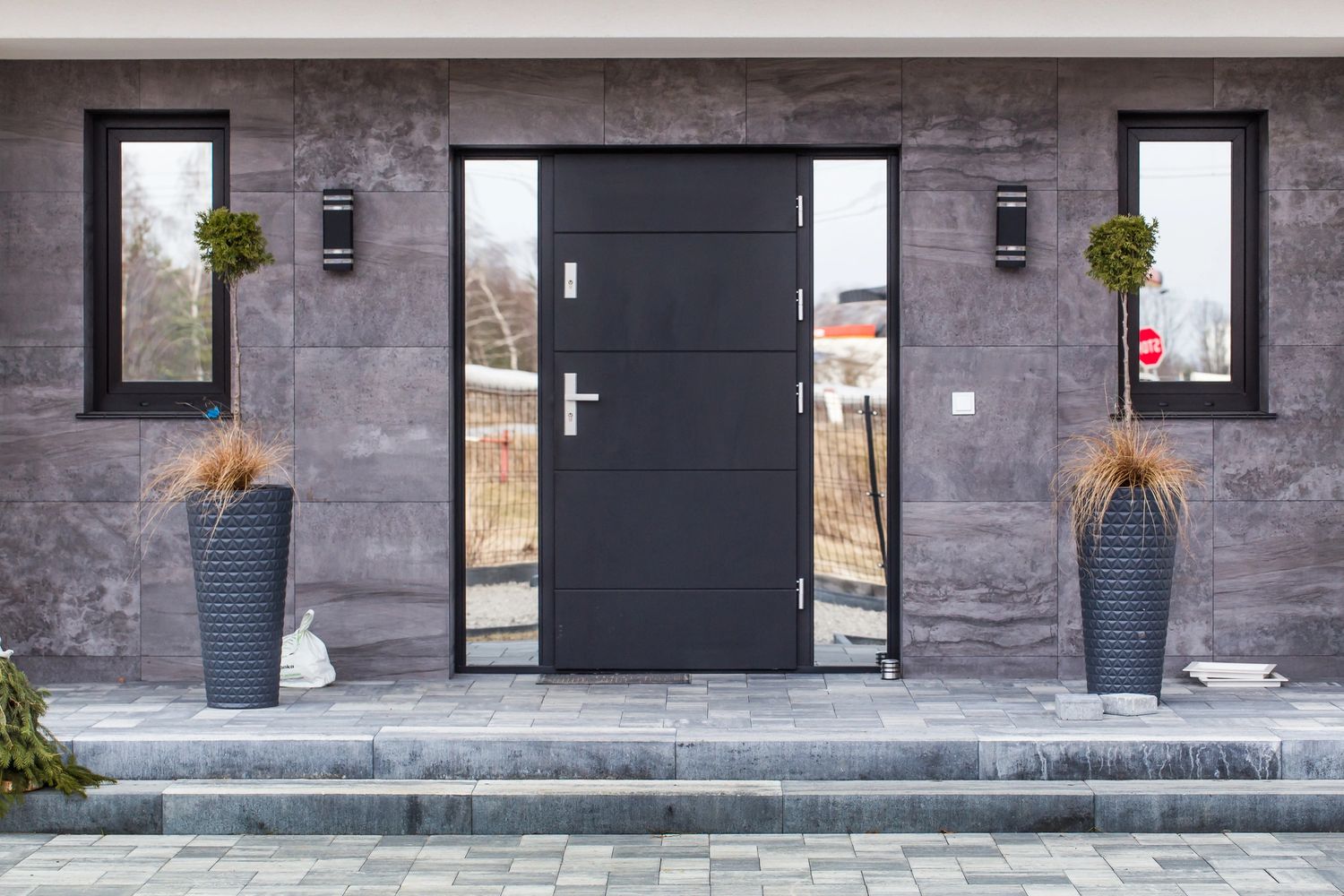 Modern entrance with a black door, two tall planters, and gray tiled walls.
