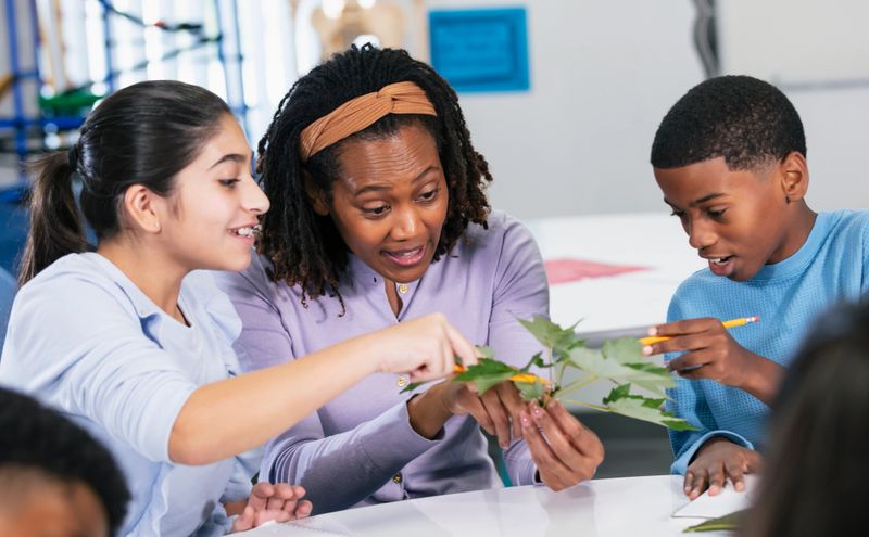 An African-American woman teaching a middle school science class. She and her students are sitting at a table, examining plant leaves and writing in notebooks. The main focus is on the teacher.