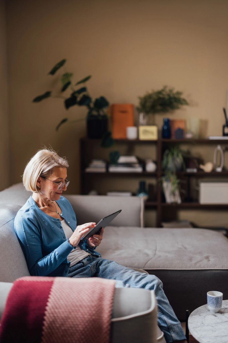 Elderly lady enjoys leisure time browsing on a tablet while sitting comfortably on her living room couch, exuding calm and contentment.