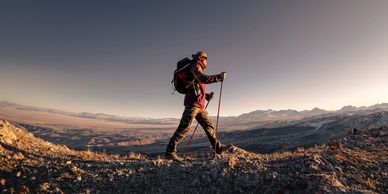 Hiker with trekking poles walking on rocky terrain at sunset.
